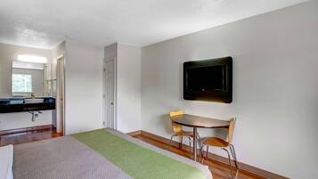 Habitación de hotel con cama, escritorio, silla, televisor montado en la pared y baño con lavamanos y espejo.