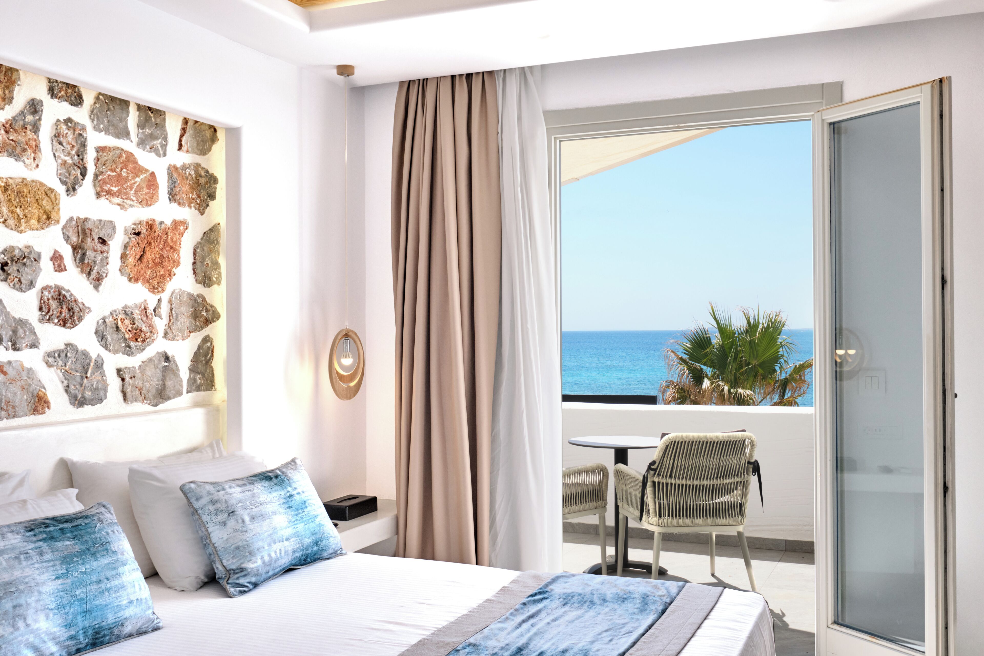 A bedroom with a stone wall, a bed with blue pillows, a balcony with a table and chair, and a view of the sea.