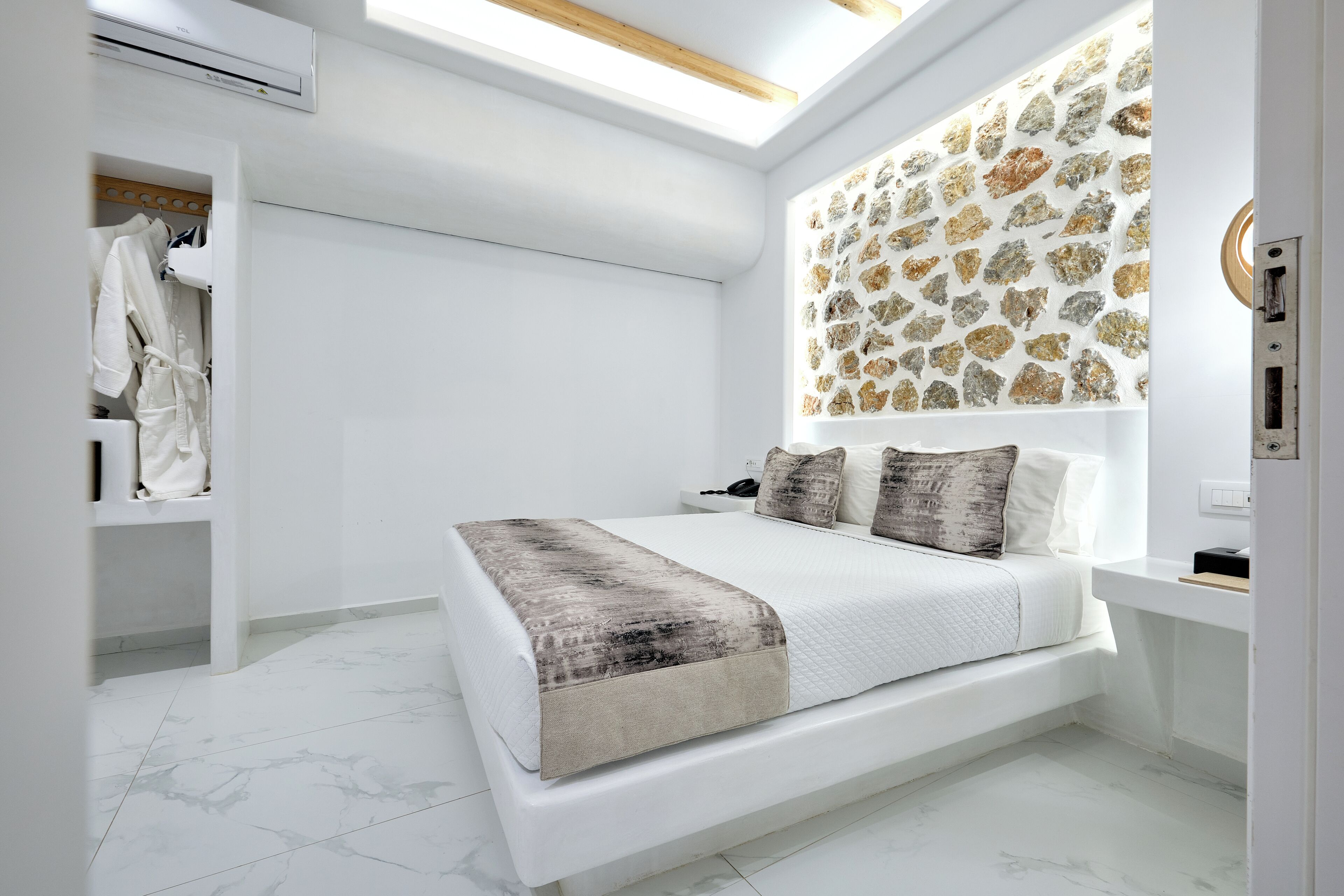A modern bedroom with a skylight, a stone-patterned wall, a bed with a white quilt, and a wardrobe with hanging robes.