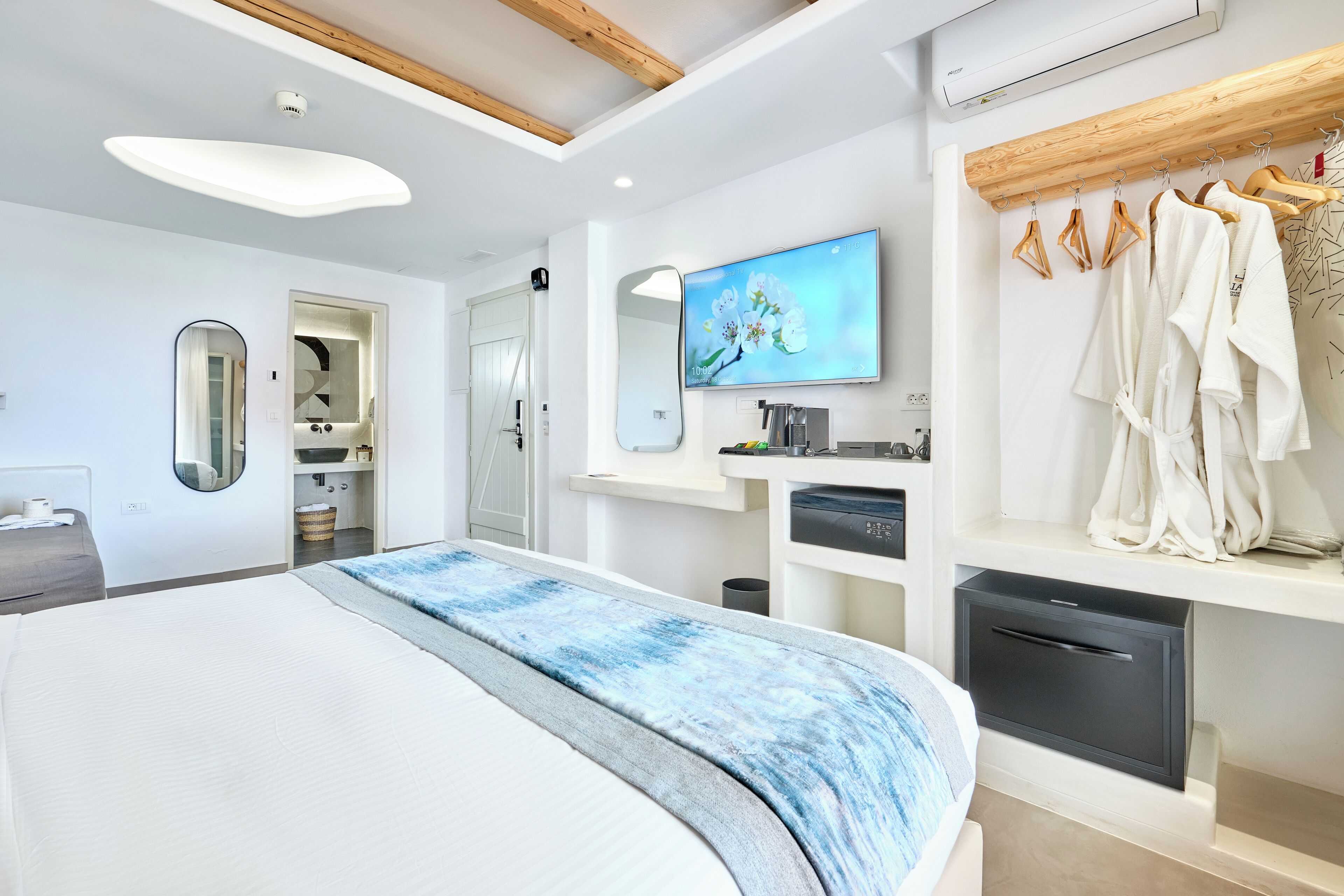 Ein modernes Hotelzimmer mit einem großen Bett, einem an der Wand montierten Fernseher, einer Holzbalkendecke und einem Kleiderständer mit Bademänteln.