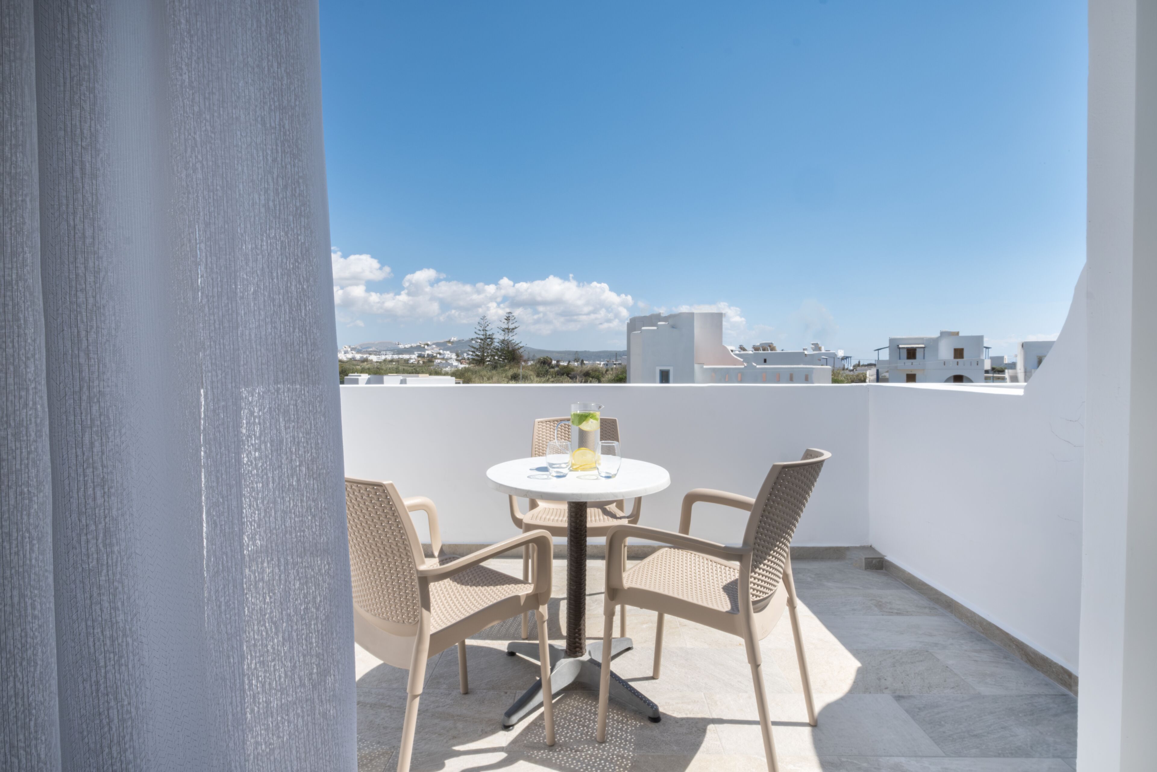 A balcony with a round table and four wicker chairs, overlooking a coastal town with white buildings and a clear sky.