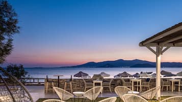 A seaside patio with wicker chairs and tables, overlooking the sea and mountains.