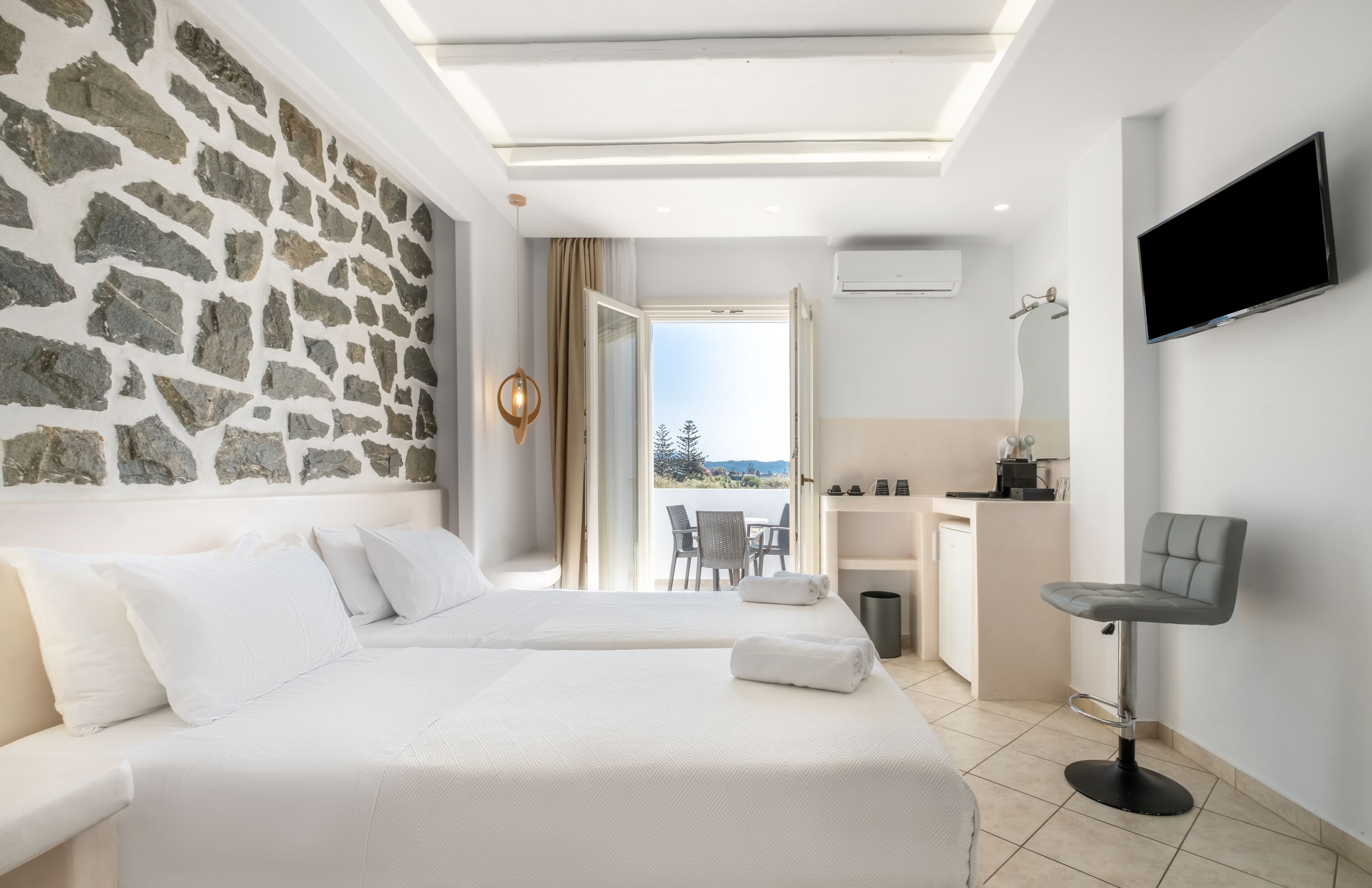 Habitación de hotel con dos camas, una pared de piedra, un balcón con vistas, un televisor y un taburete de bar.
