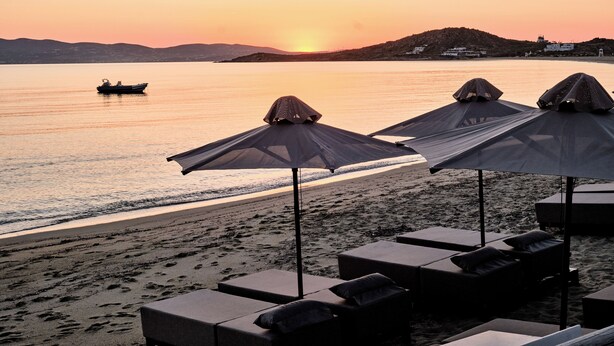A beach with lounge chairs and umbrellas, a boat on the water, and a sunset.