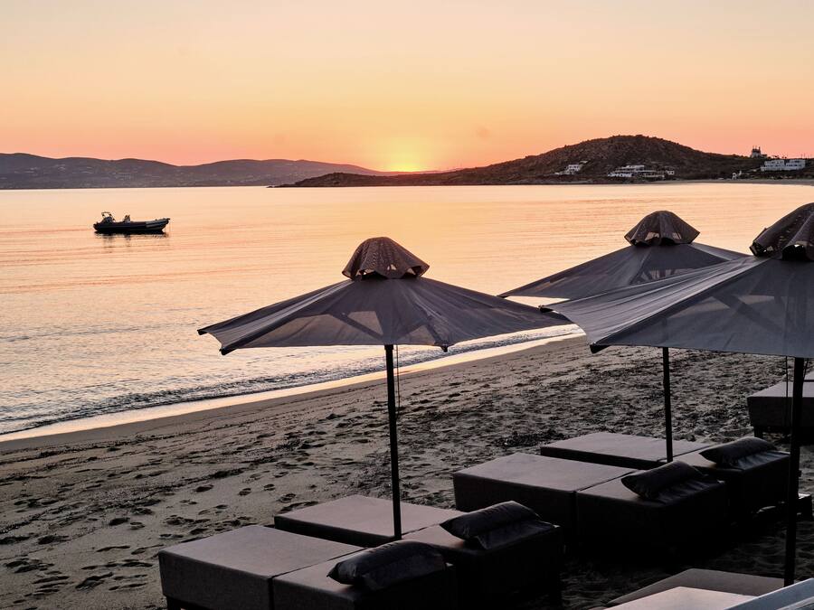 Una playa con sillas de descanso y sombrillas, un barco en el agua y una puesta de sol.