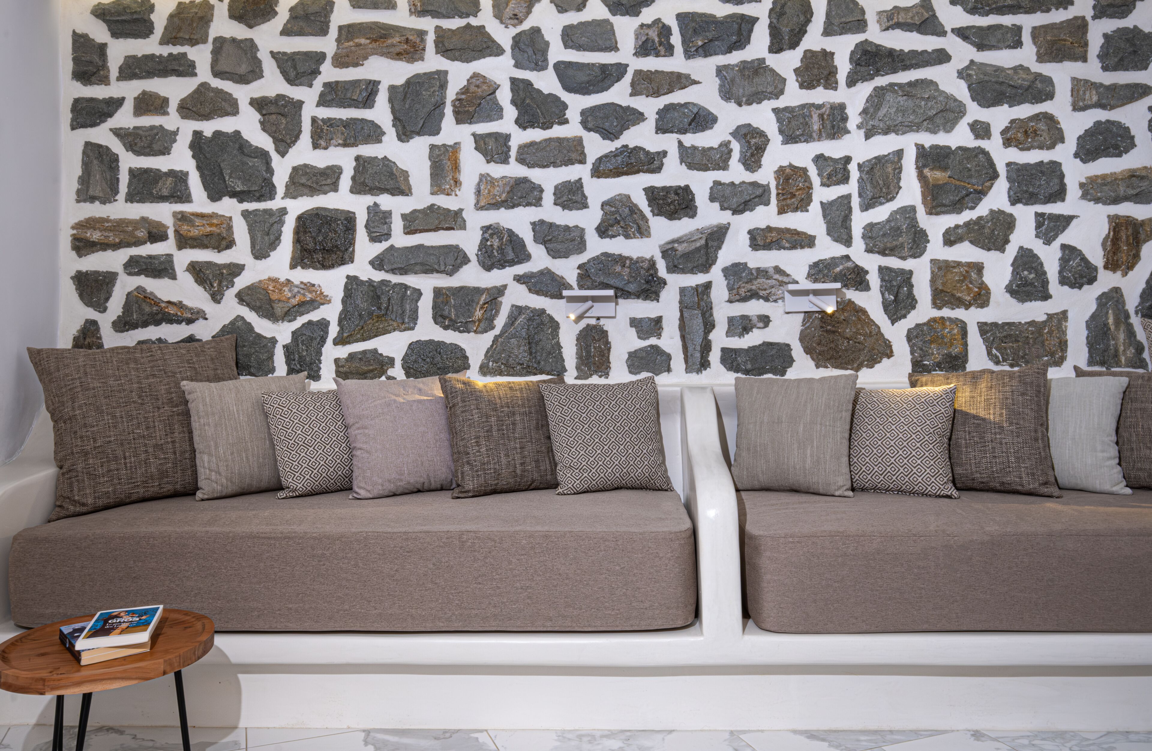 Una sala de estar moderna con una pared de piedra, un sofá beige con cojines estampados y una pequeña mesa auxiliar de madera con un libro.