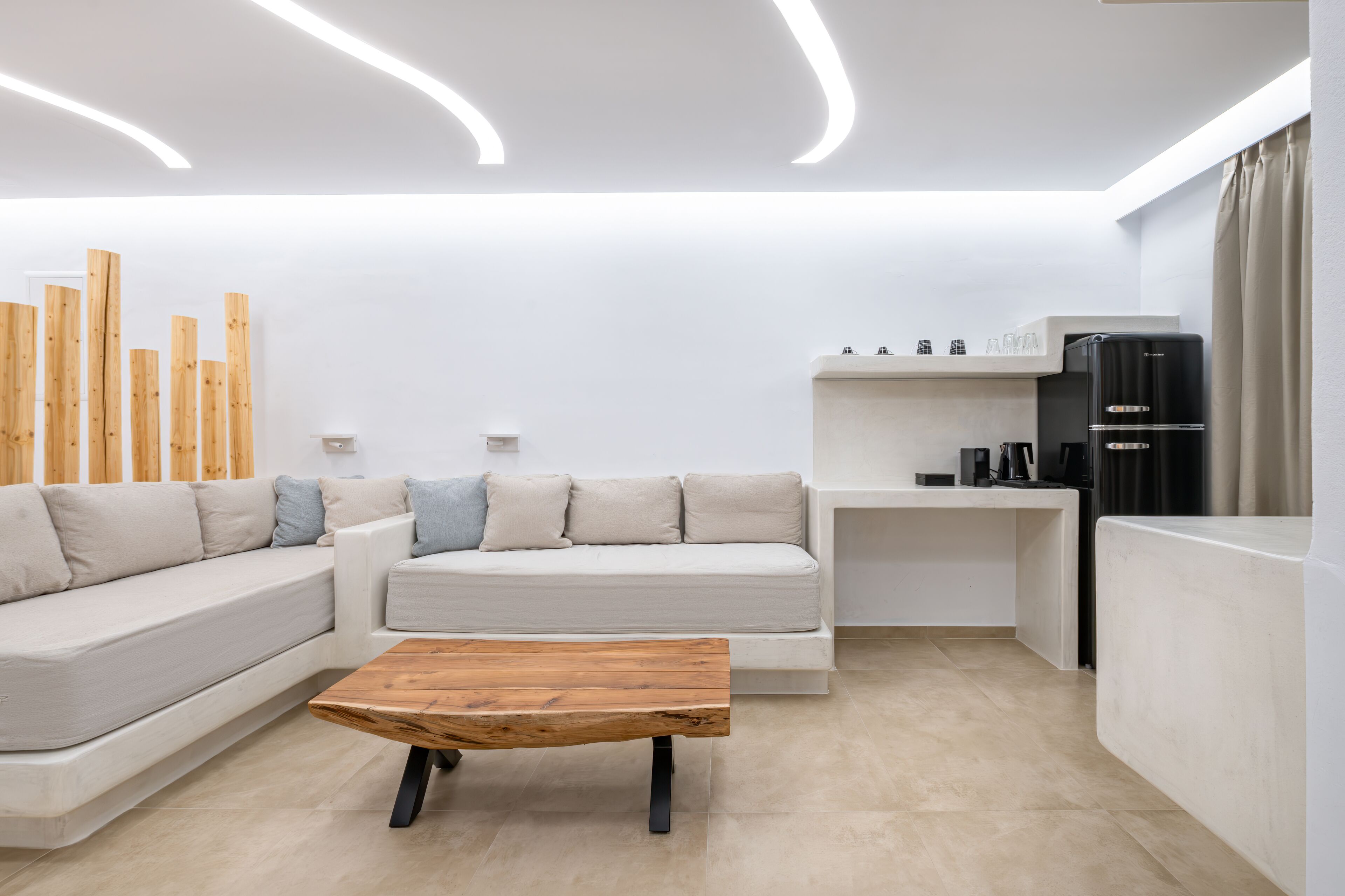 A modern living room with a sectional sofa, a wooden coffee table, a built-in kitchen area with a black refrigerator, and a minimalist design.