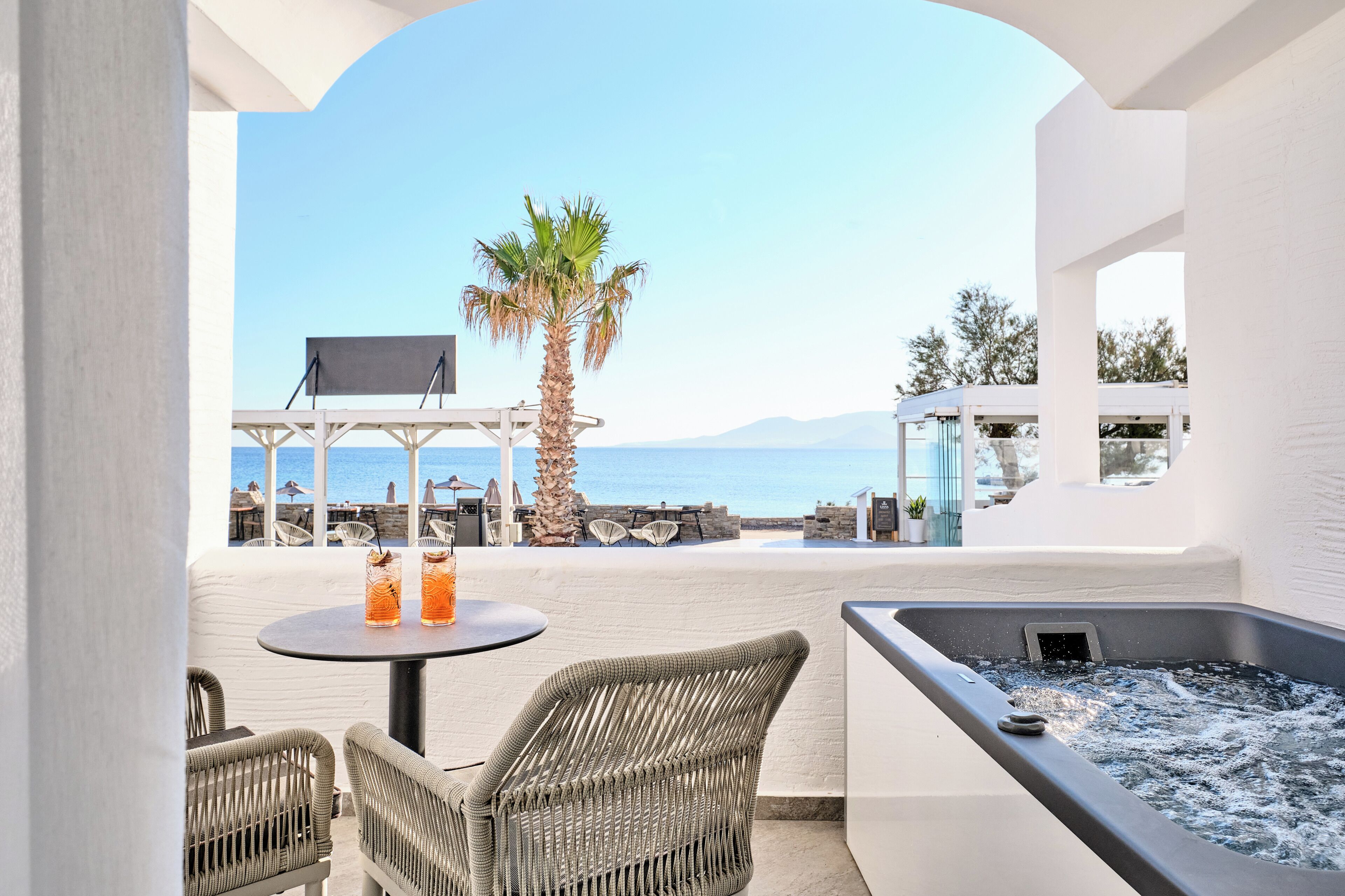A balcony with wicker chairs, a table with drinks, a hot tub, and a view of the sea and palm tree.