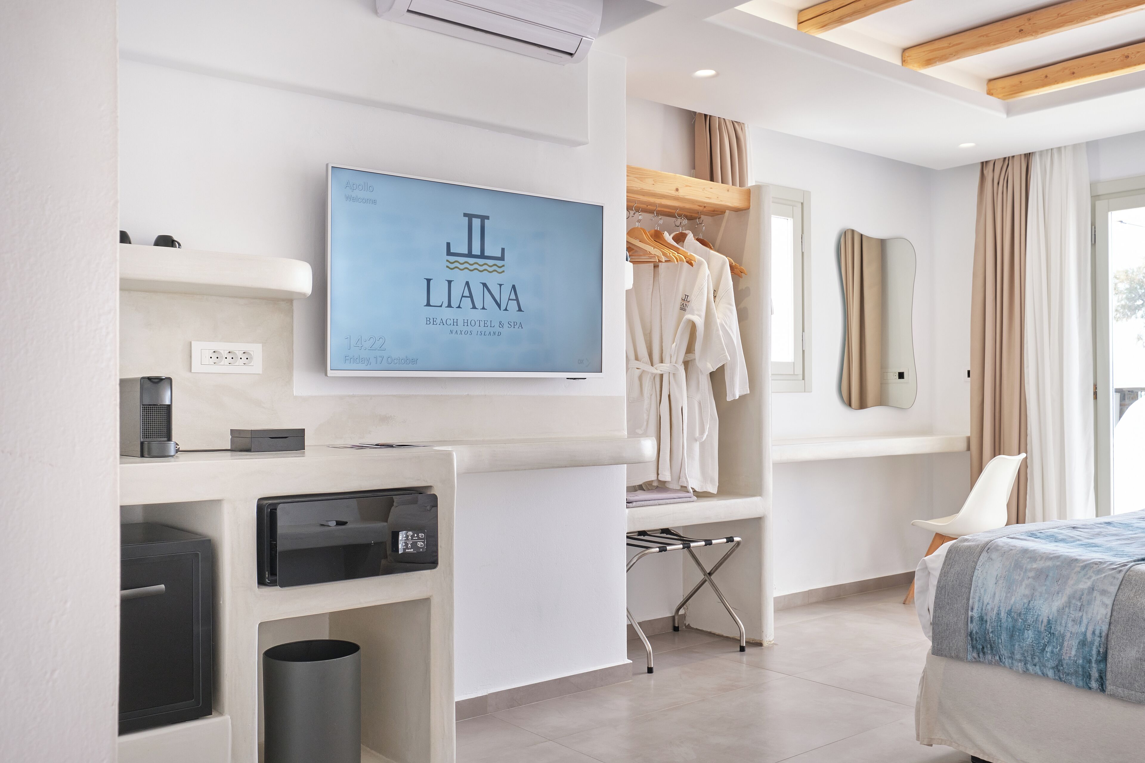 A modern hotel room with a flat-screen TV displaying the hotel's name, a minibar, a desk with a coffee machine, a hanging robe, a mirror, and a bed with a blue and white bedspread.