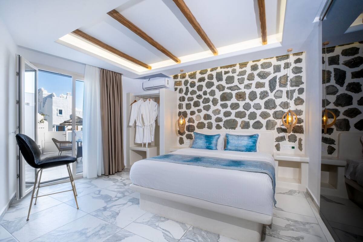 A modern bedroom with a stone wall, a bed with blue pillows, a chair, and a view of the city.