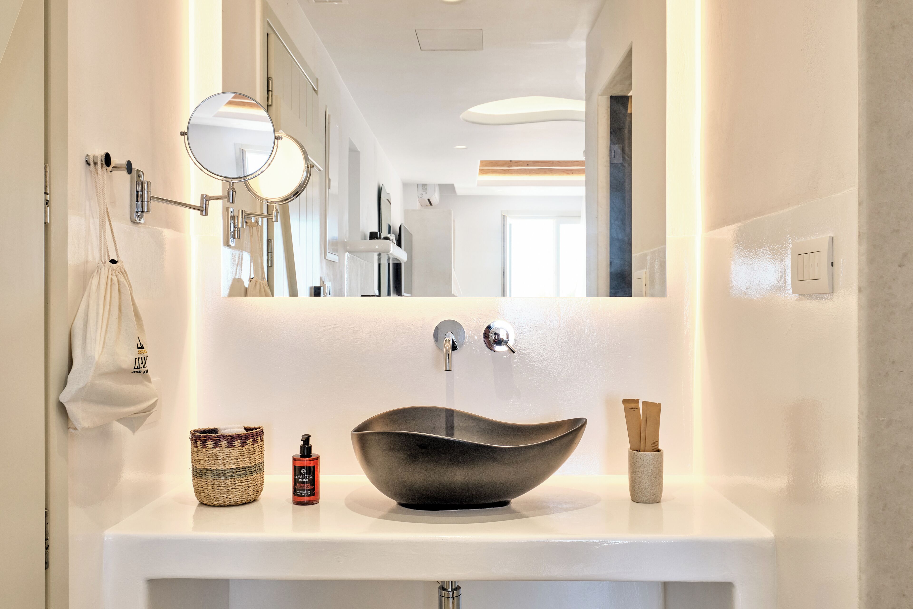 Un baño moderno con un lavabo blanco, un espejo redondo, una toalla colgada, una cesta tejida y un pequeño recipiente con cepillos de dientes.