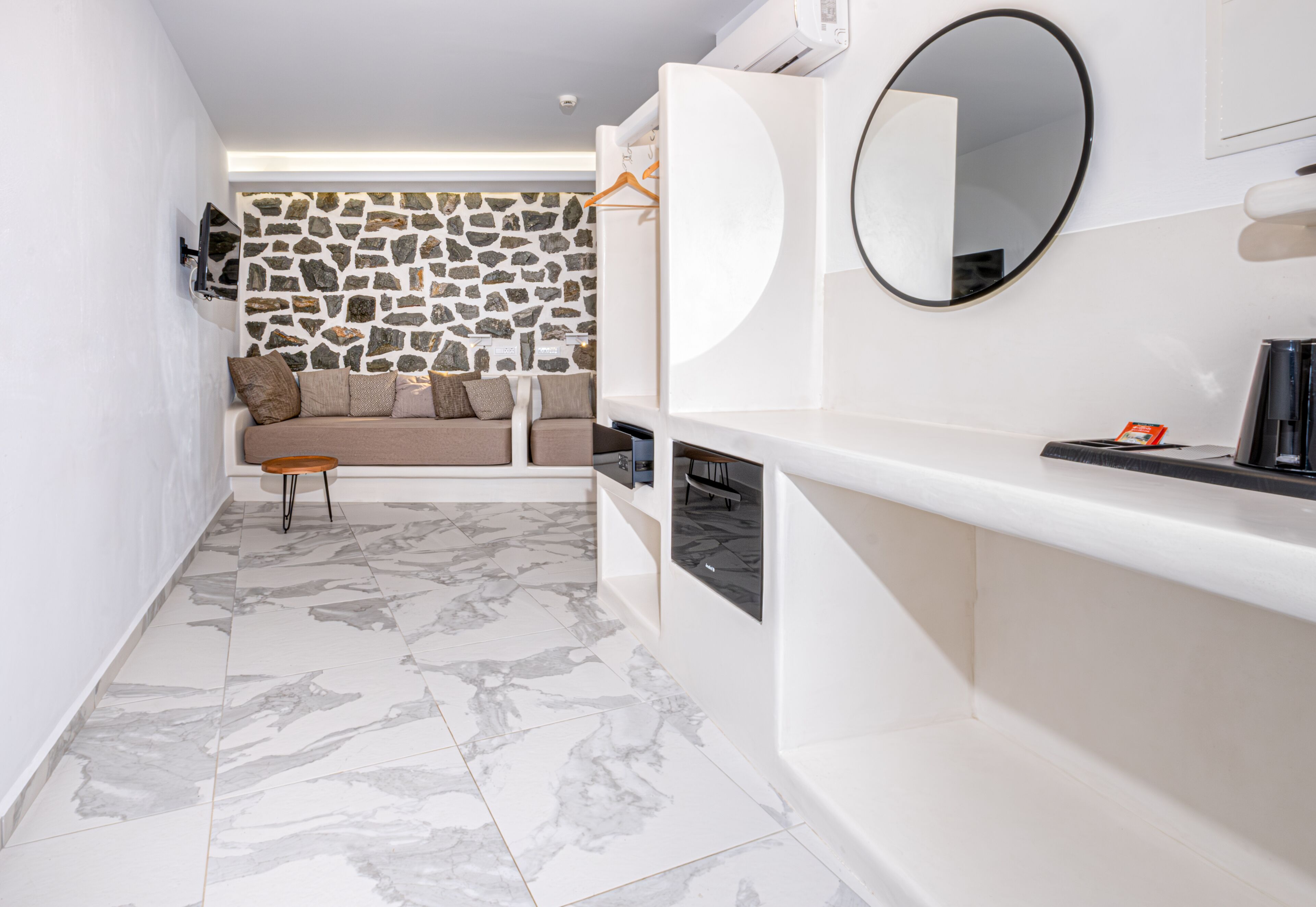 Una habitación de hotel moderna con pared de piedra, un sofá con cojines, una mesita, un espejo y una cafetera.