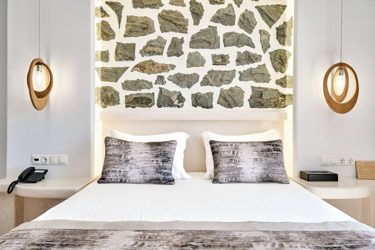A hotel room with a stone wall, a bed with white bedding, two pillows, a bedside table with a phone, and a wall-mounted light fixture.