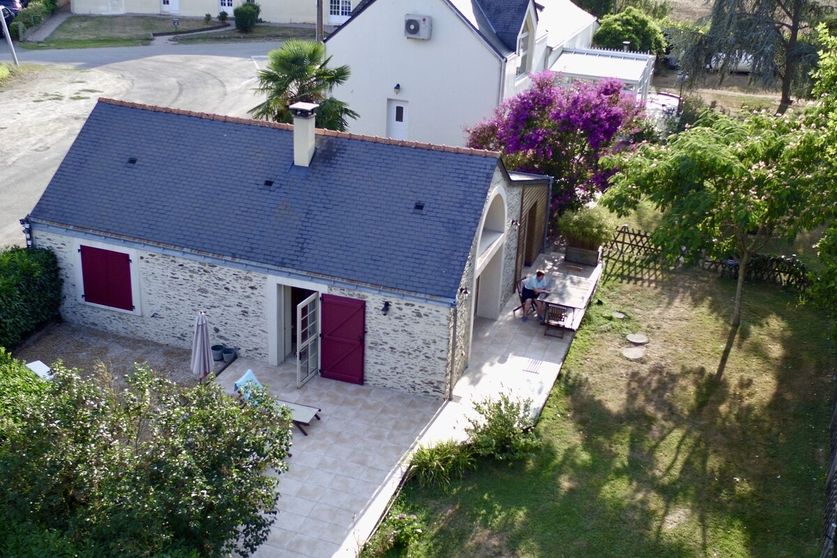 Un bâtiment à deux étages, doté d’un toit en tuiles, d’un patio avec une table et des chaises, et d’un jardin agrémenté de plantes à fleurs.