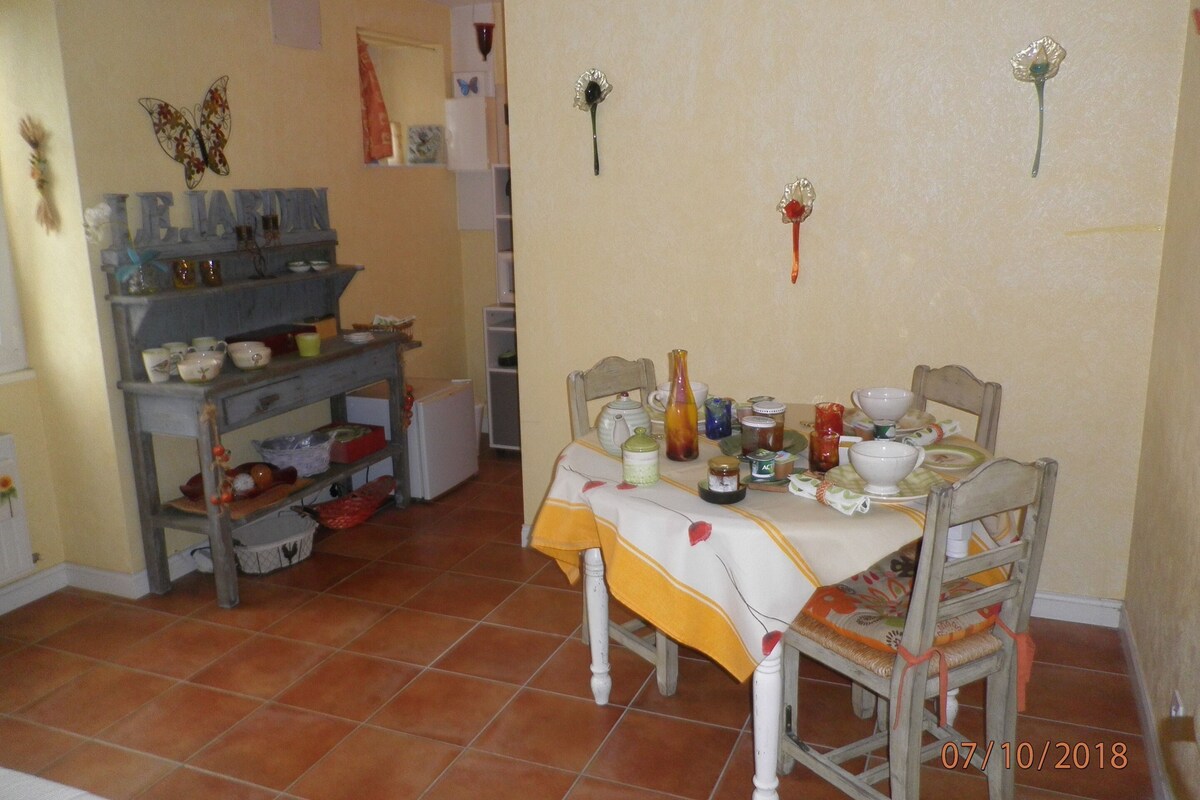 Un coin repas avec une table dressée pour deux personnes, entourée de chaises. Une étagère contenant divers ustensiles de cuisine est visible sur la gauche.