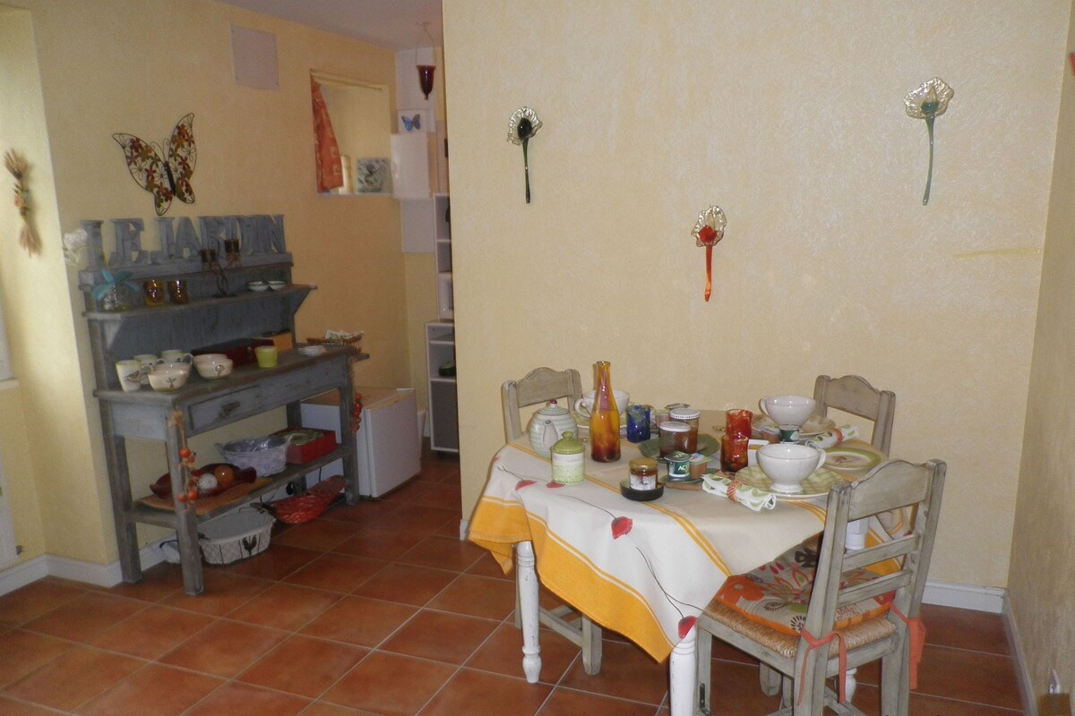 Un coin repas avec une table dressée pour deux personnes, entourée de chaises. Une étagère contenant divers ustensiles de cuisine est visible sur la gauche.