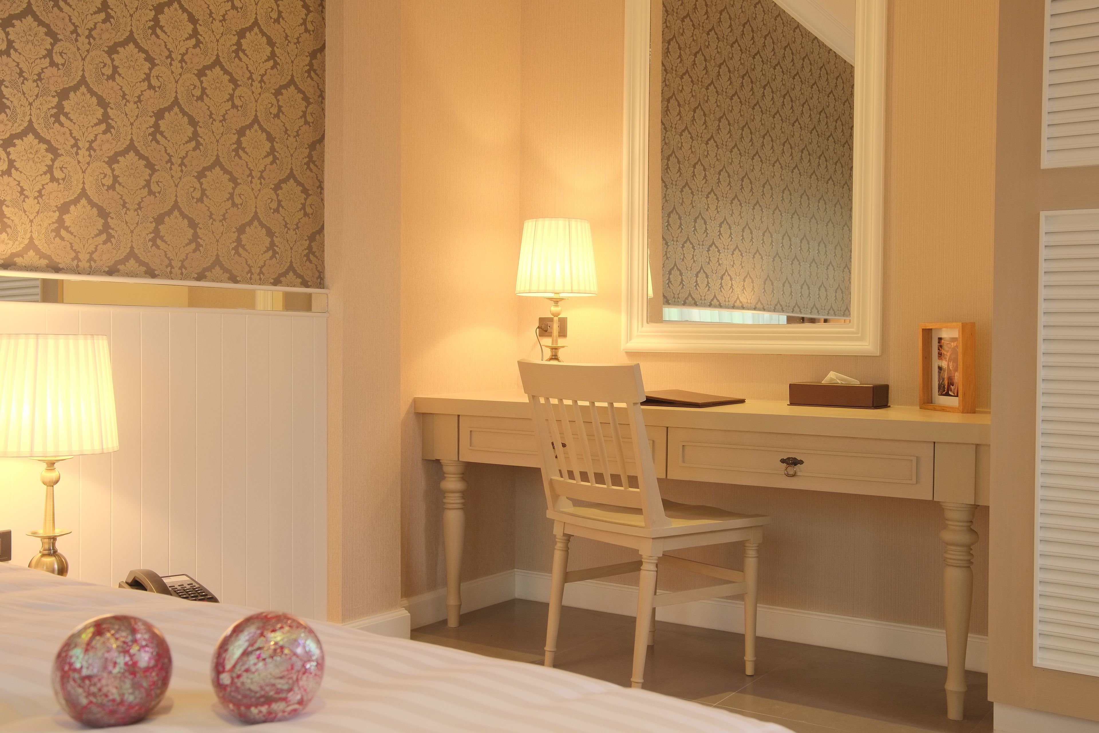 A hotel room with a bed, a desk with a chair, a lamp, a telephone, and a window with a patterned curtain.