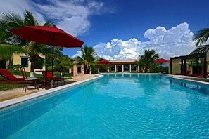 Une piscine bordée de parasols rouges, de palmiers et surplombée d’un ciel bleu azur.