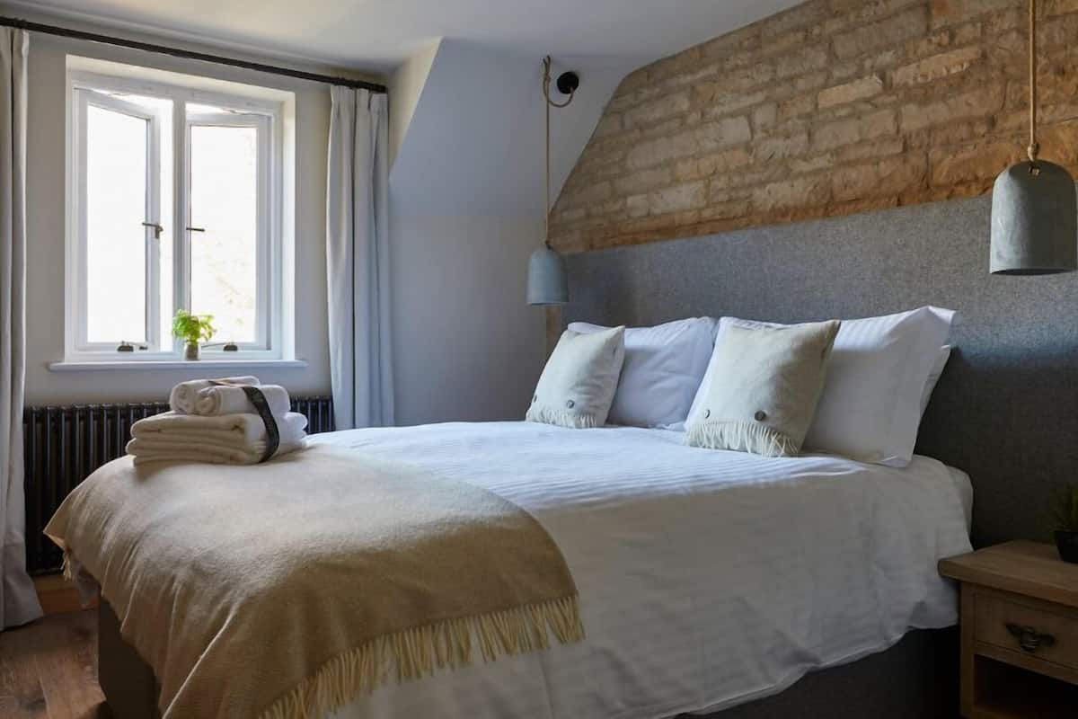Un lit bien fait, avec une couverture et des oreillers de couleur beige, une fenêtre avec des rideaux, un radiateur et un mur en briques.