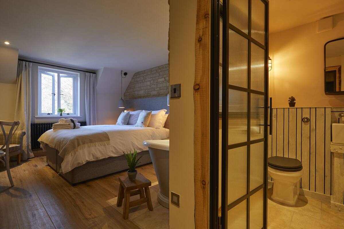 Une chambre à coucher avec un lit, une fenêtre, un radiateur et un petit tabouret sur lequel est posée une plante.