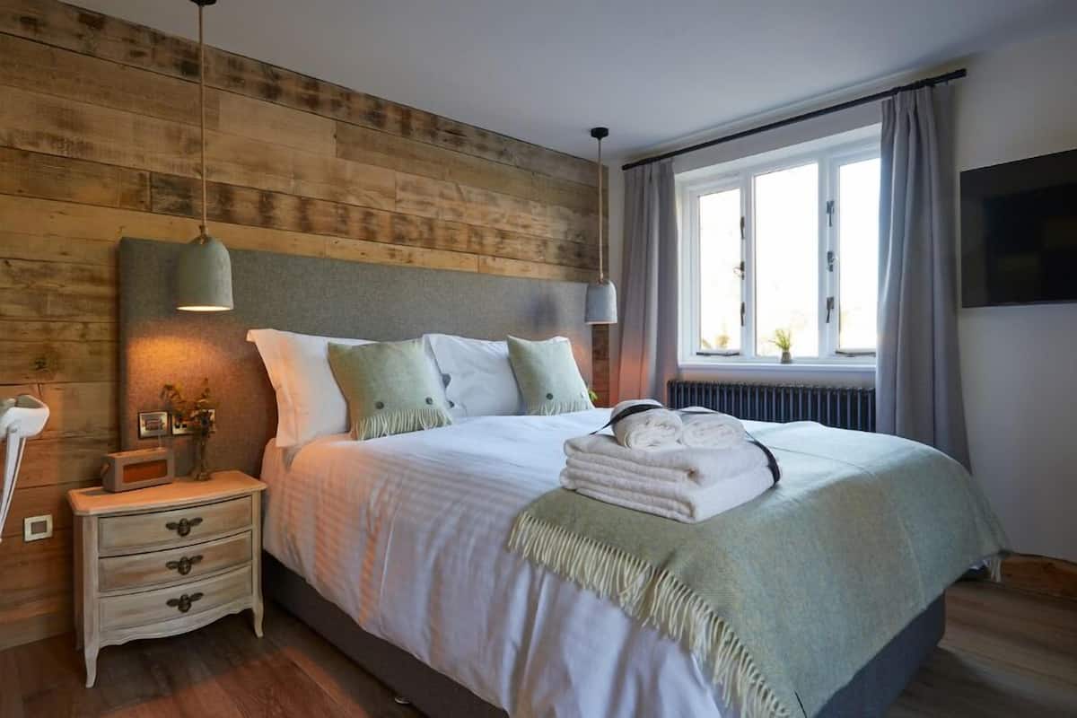 Une chambre à coucher avec une tête de lit en bois, un lit recouvert de draps blancs, une table de chevet avec une lampe et une fenêtre avec des rideaux.