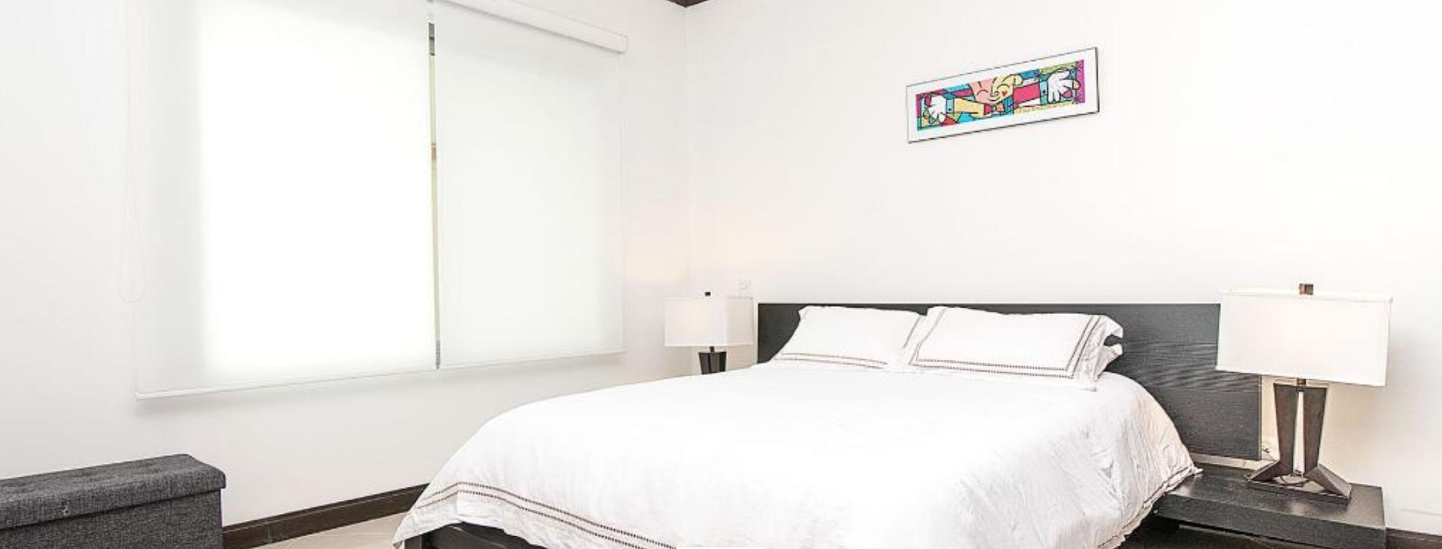 Ein Hotelzimmer mit einem großen Bett, zwei Nachttischlampen, einer grauen Bank und einem farbenfrohen Kunstwerk an der Wand.