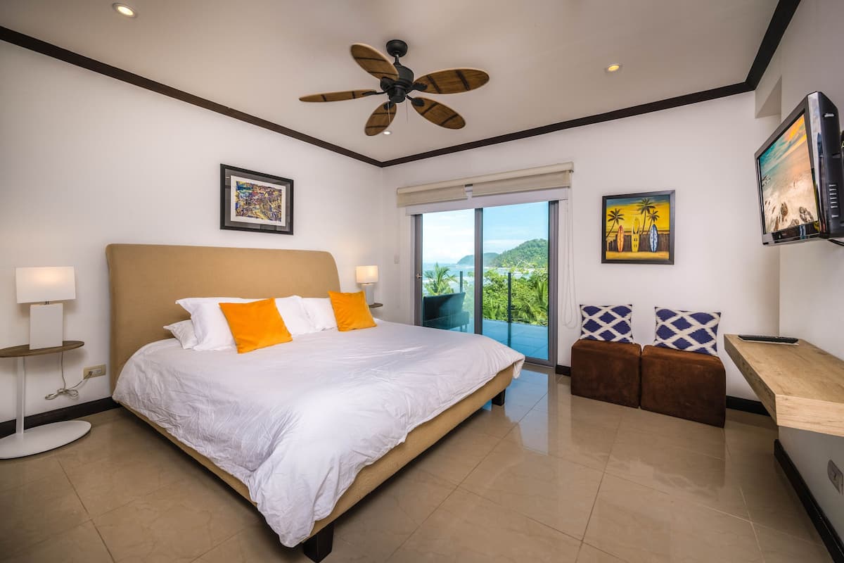 A modern hotel room with a large bed, a ceiling fan, a TV, and a balcony with a view of the mountains.