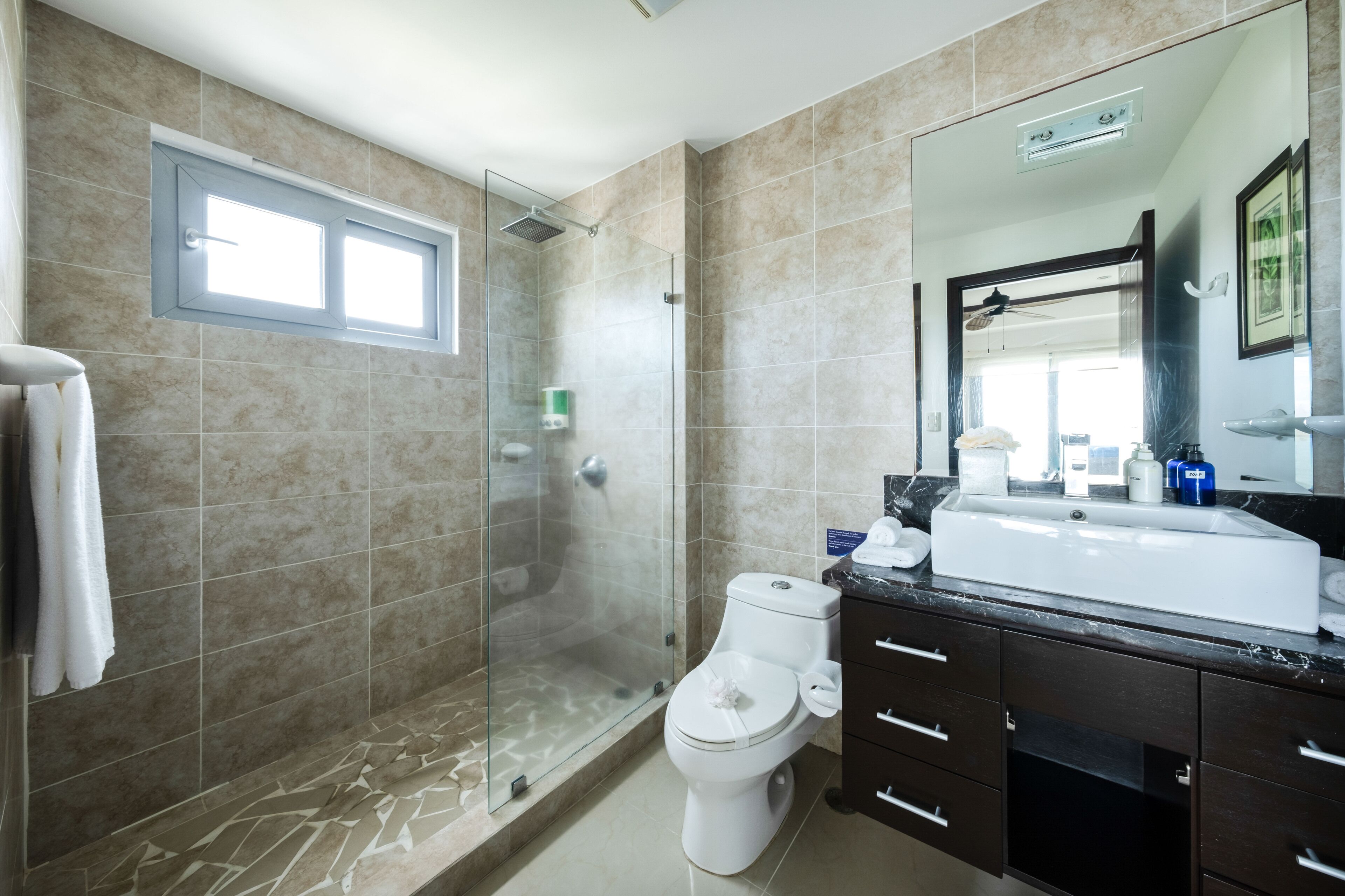 A bathroom with a glass-enclosed shower, a toilet, a sink with a mirror above it, and a window above the shower.
