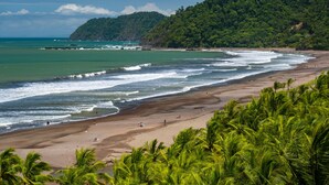 A beach with clear waters, a sandy shore, and lush greenery in the background. - Best in Jaco Diamante del Sol Condos (Jacó)