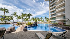 A swimming pool with palm trees, lounge chairs, and a building with balconies. - Best in Jaco Diamante del Sol Condos (Jacó)