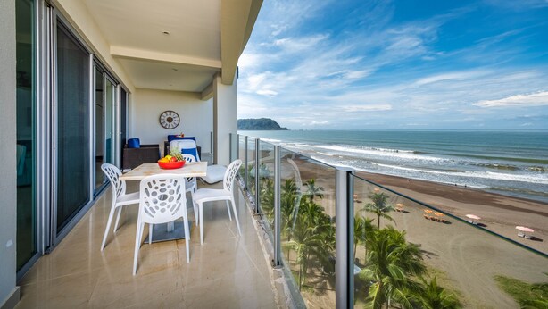 A balcony with a table and chairs, overlooking a beach with umbrellas and palm trees. - Best in Jaco Diamante del Sol Condos (Jacó)