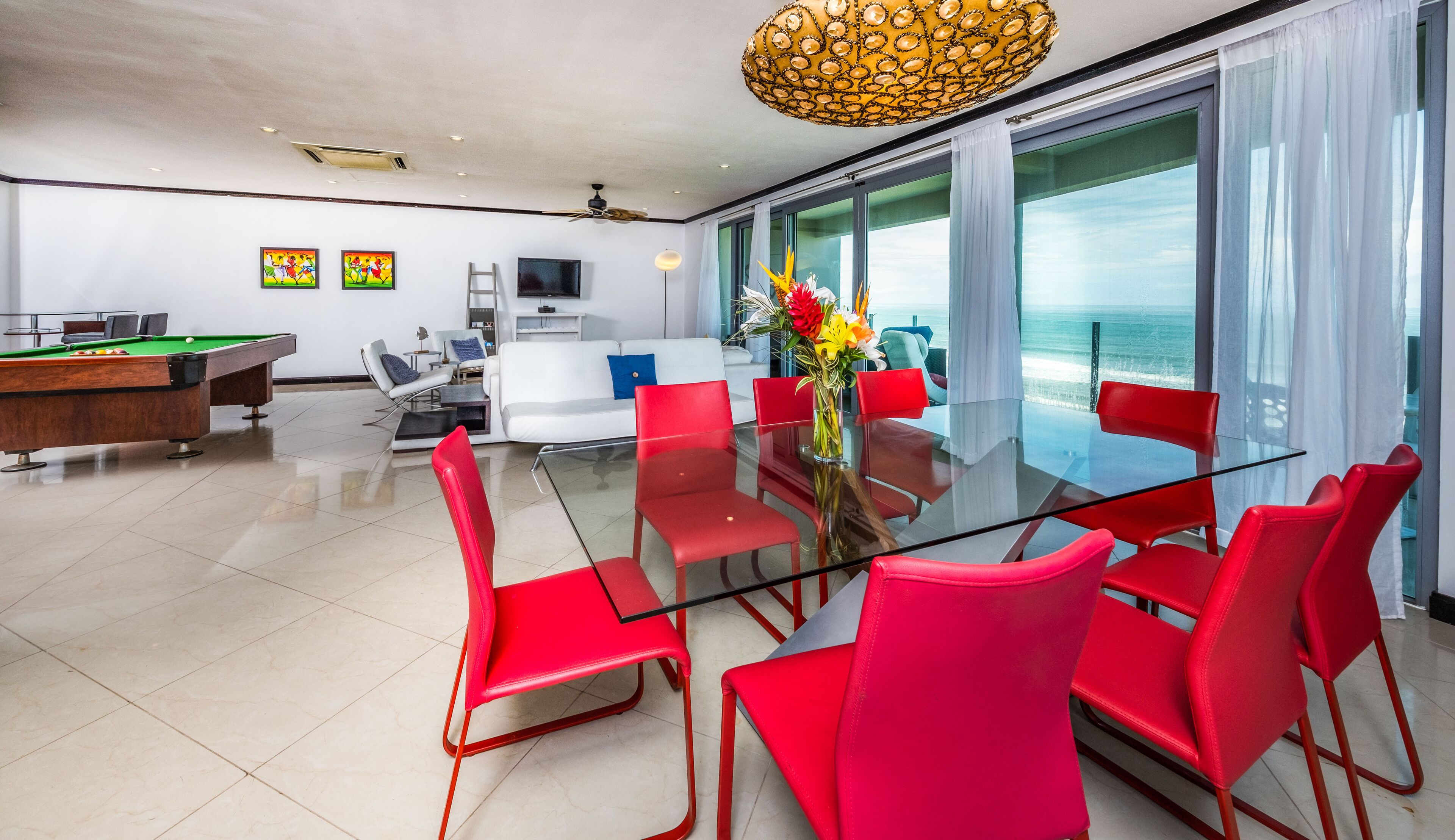A modern living room with a glass table, red chairs, a pool table, and a view of the ocean.