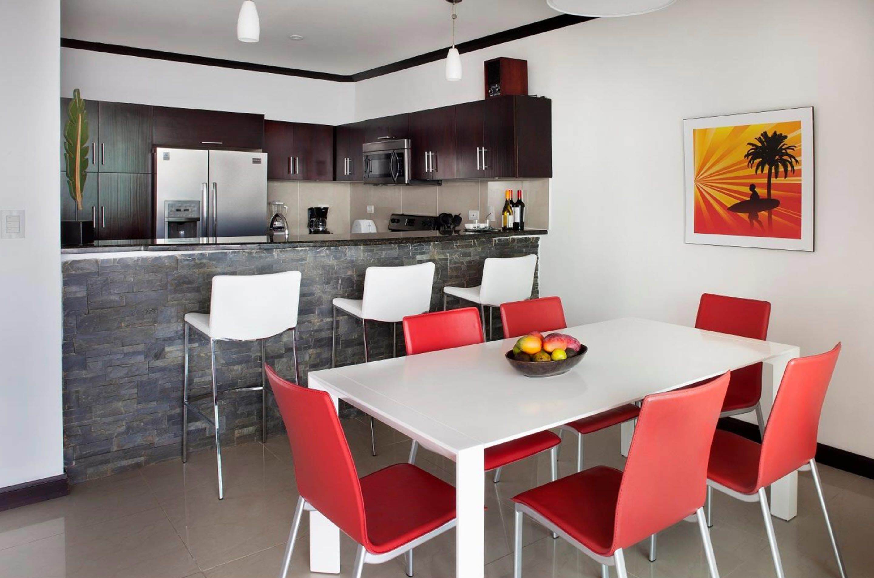 Una cocina moderna con armarios oscuros, electrodomésticos de acero inoxidable y una encimera de piedra. Hay un comedor con una mesa blanca y sillas rojas.