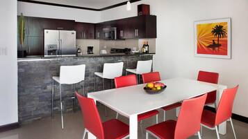 Una cocina moderna con armarios oscuros, electrodomésticos de acero inoxidable y una encimera de piedra. Hay un comedor con una mesa blanca y sillas rojas.