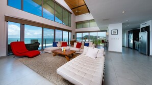 A spacious living room with a large sofa, a red chair, and a coffee table. There are large windows offering a view of the ocean. - Best in Jaco Diamante del Sol Condos (Jacó)