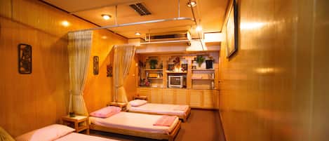 A hotel room with three single beds, a small table with a lamp, a television, and a shelf with decorative items.