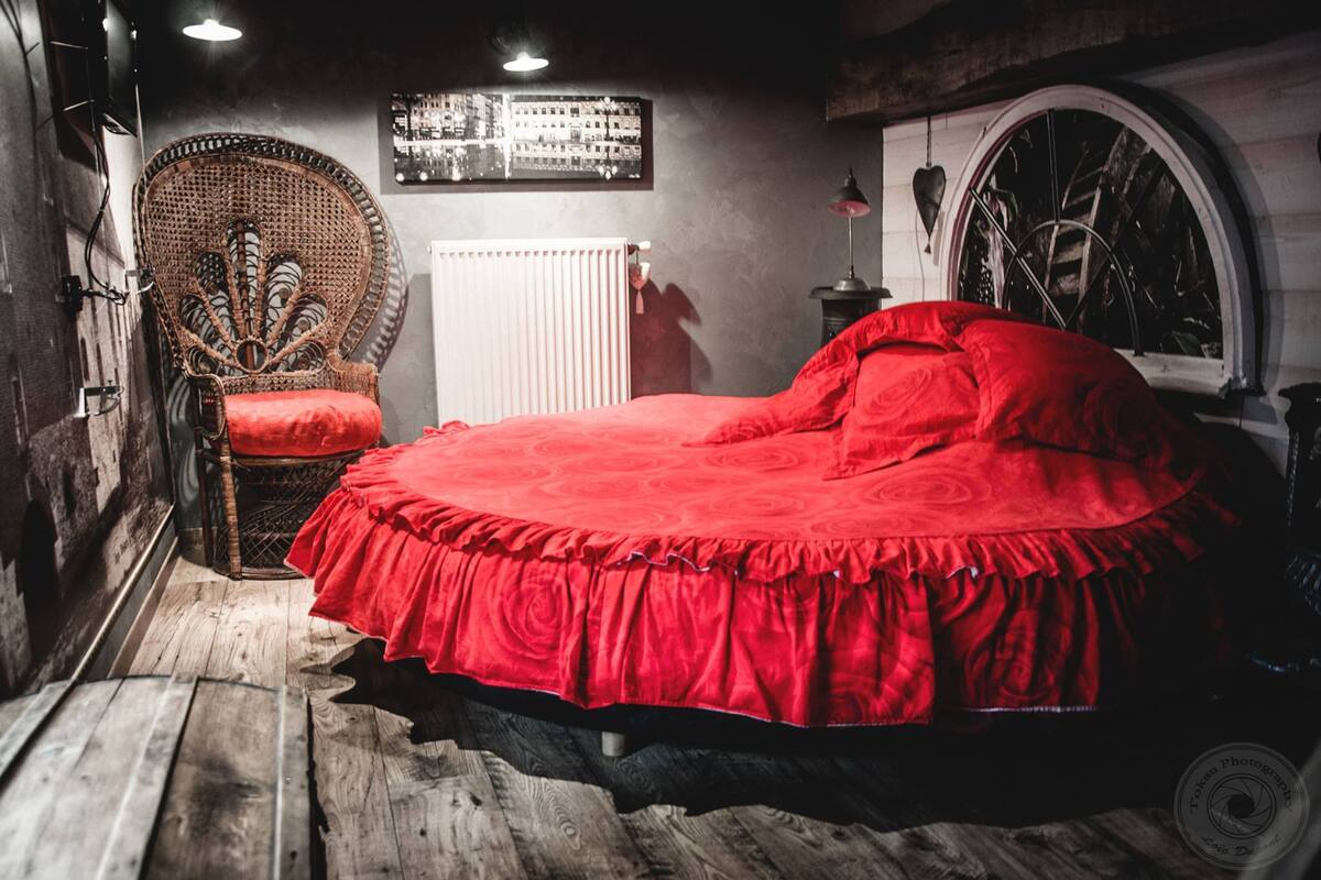 Una habitación con piso de madera, una cama roja con cubrecama fruncido, una silla de mimbre con cojín rojo, un radiador y un cuadro colgado en la pared.