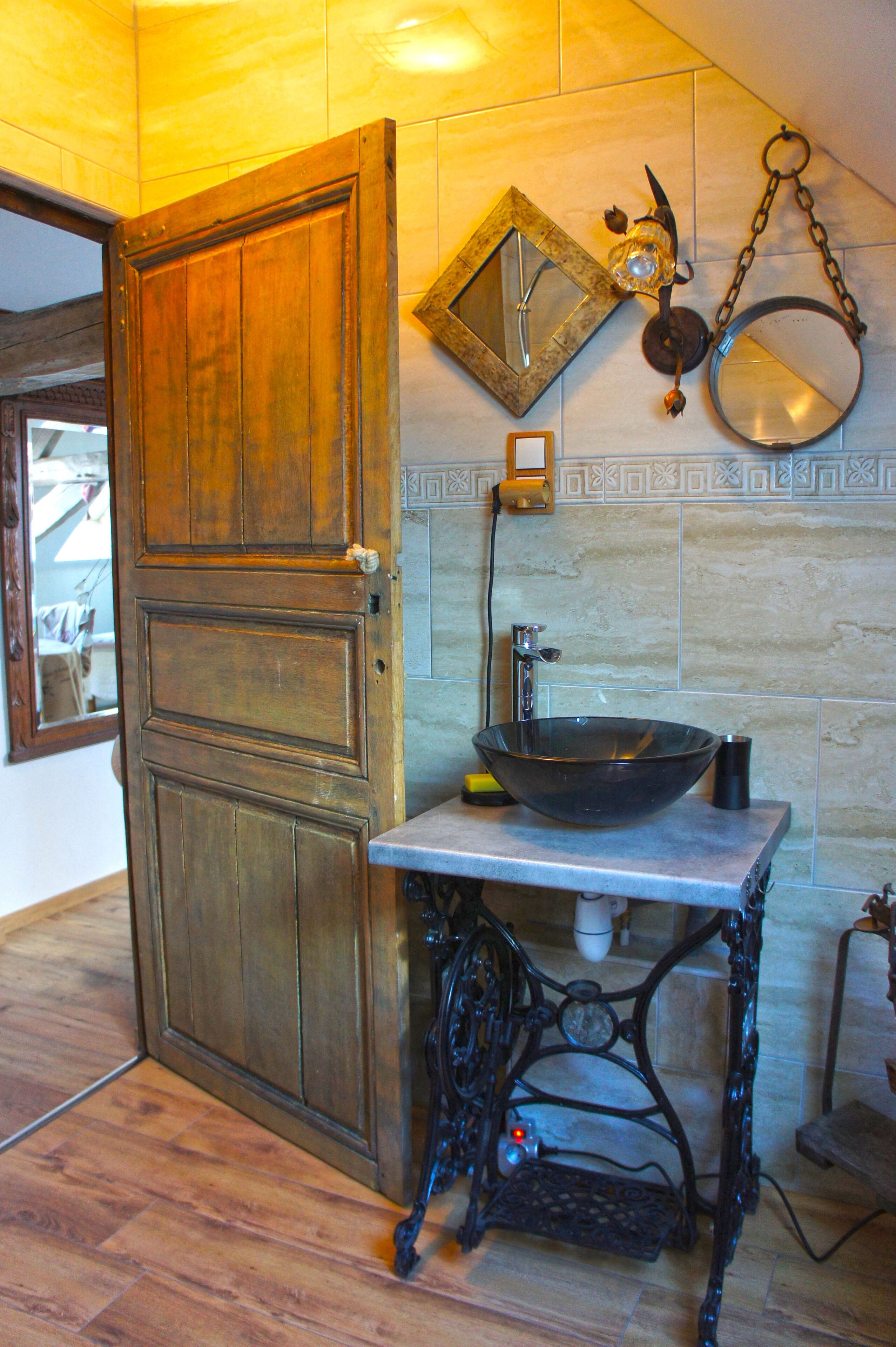A bathroom with a wooden door, a sink with a basin, a mirror, and a wall-mounted light fixture.