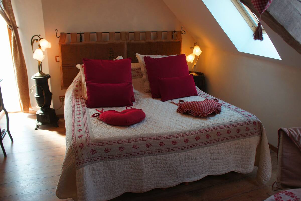 Un dormitorio acogedor con techo inclinado, cama con almohadas rojas, colcha acolchada y dos lámparas fijadas a la pared.