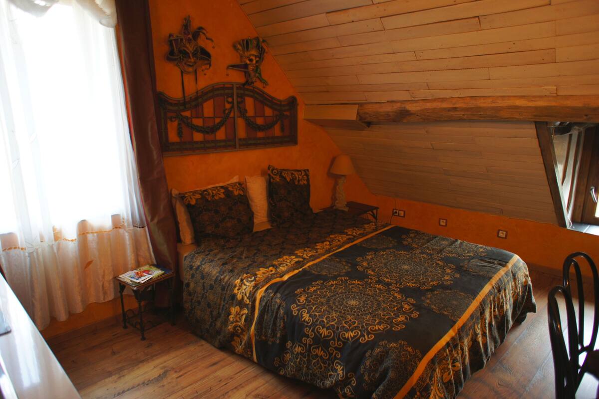 Un dormitorio con techo de madera, una cama con colcha estampada, una mesita de noche, una silla y una ventana con cortinas.