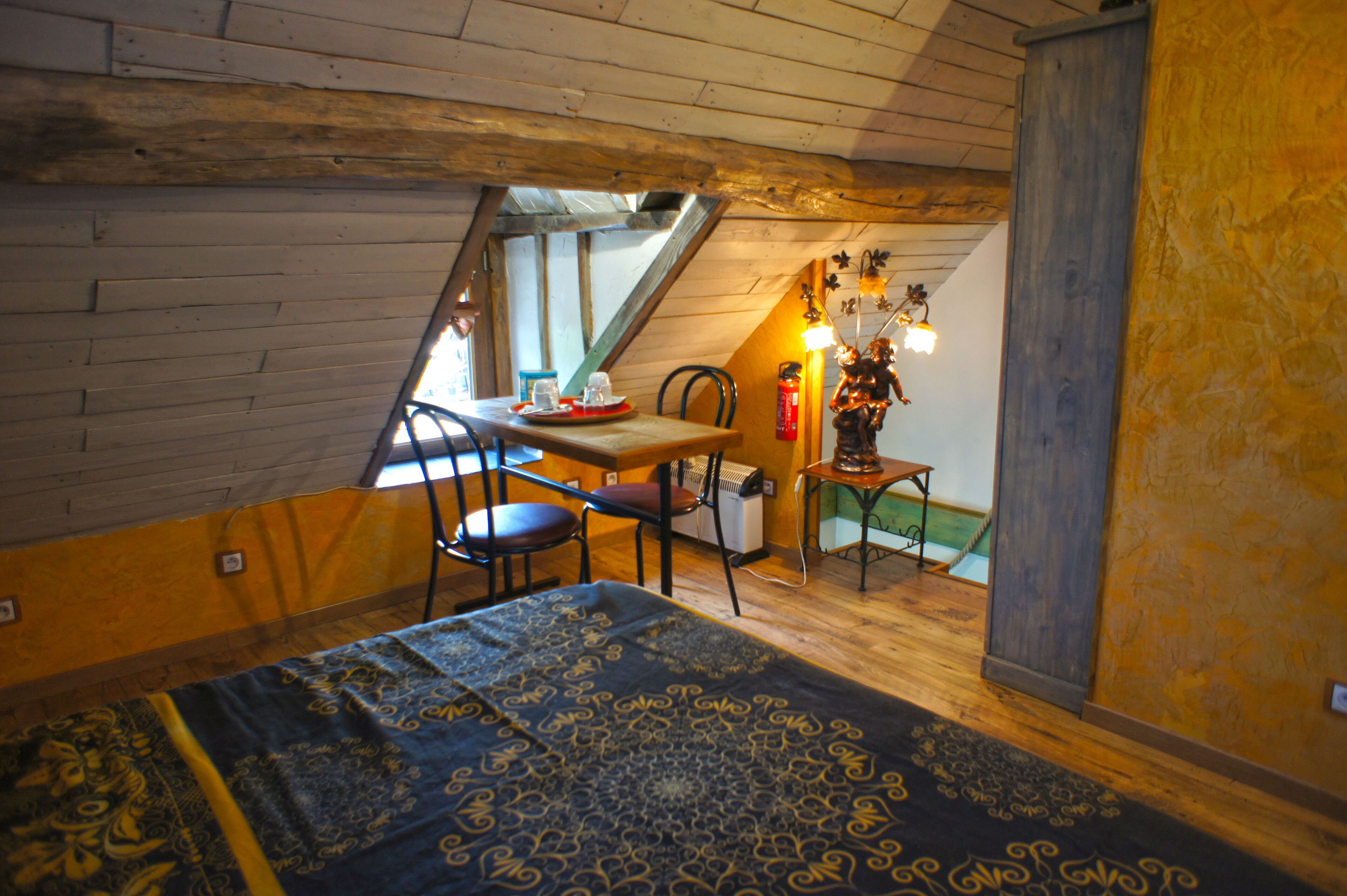 A room with a sloped ceiling, wooden walls, a bed with a patterned bedspread, a desk with a chair, and a small table with a statue.