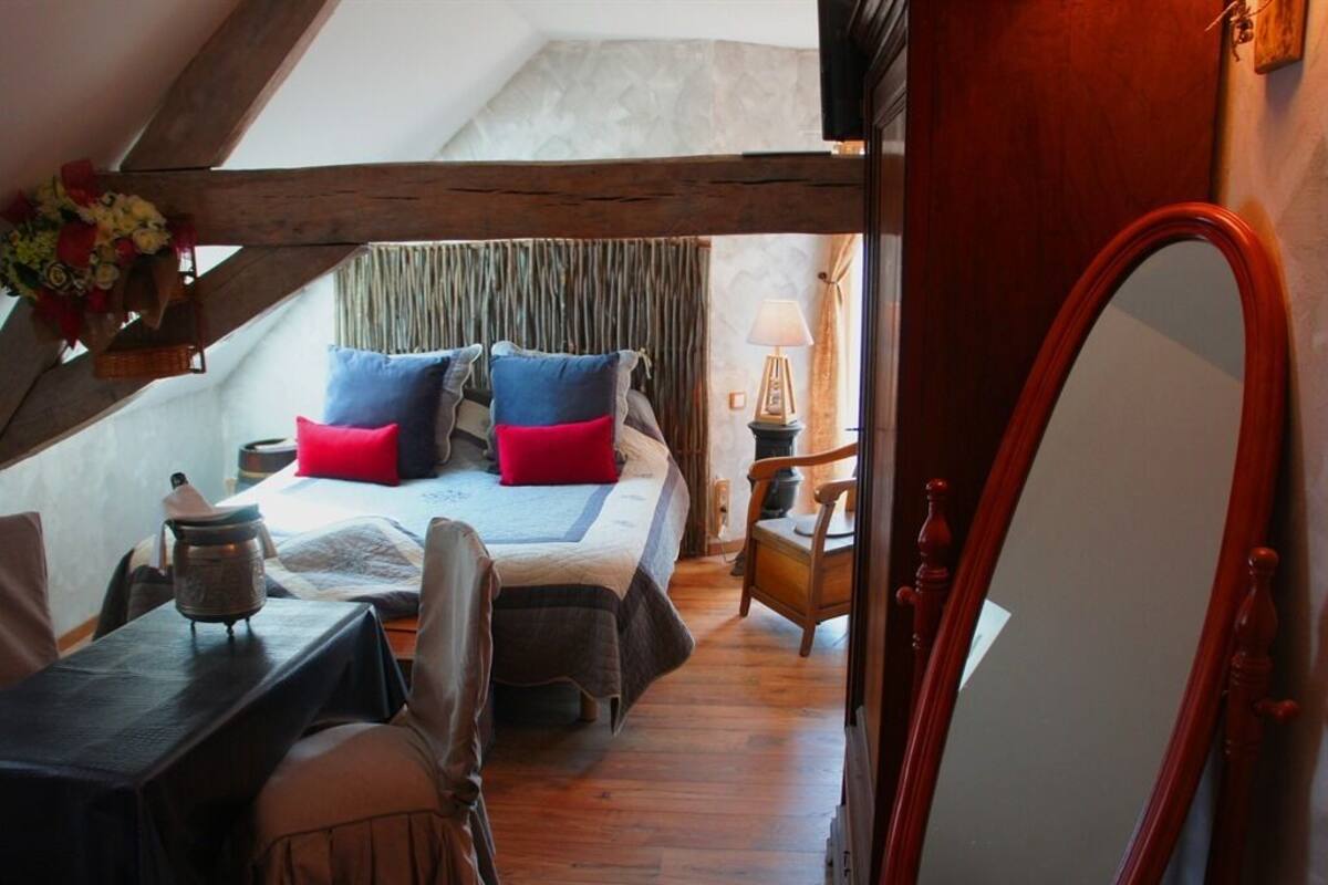 Un dormitorio acogedor en el ático con techo de madera, una cama con muchas almohadas, una mesita con una tetera y un espejo.