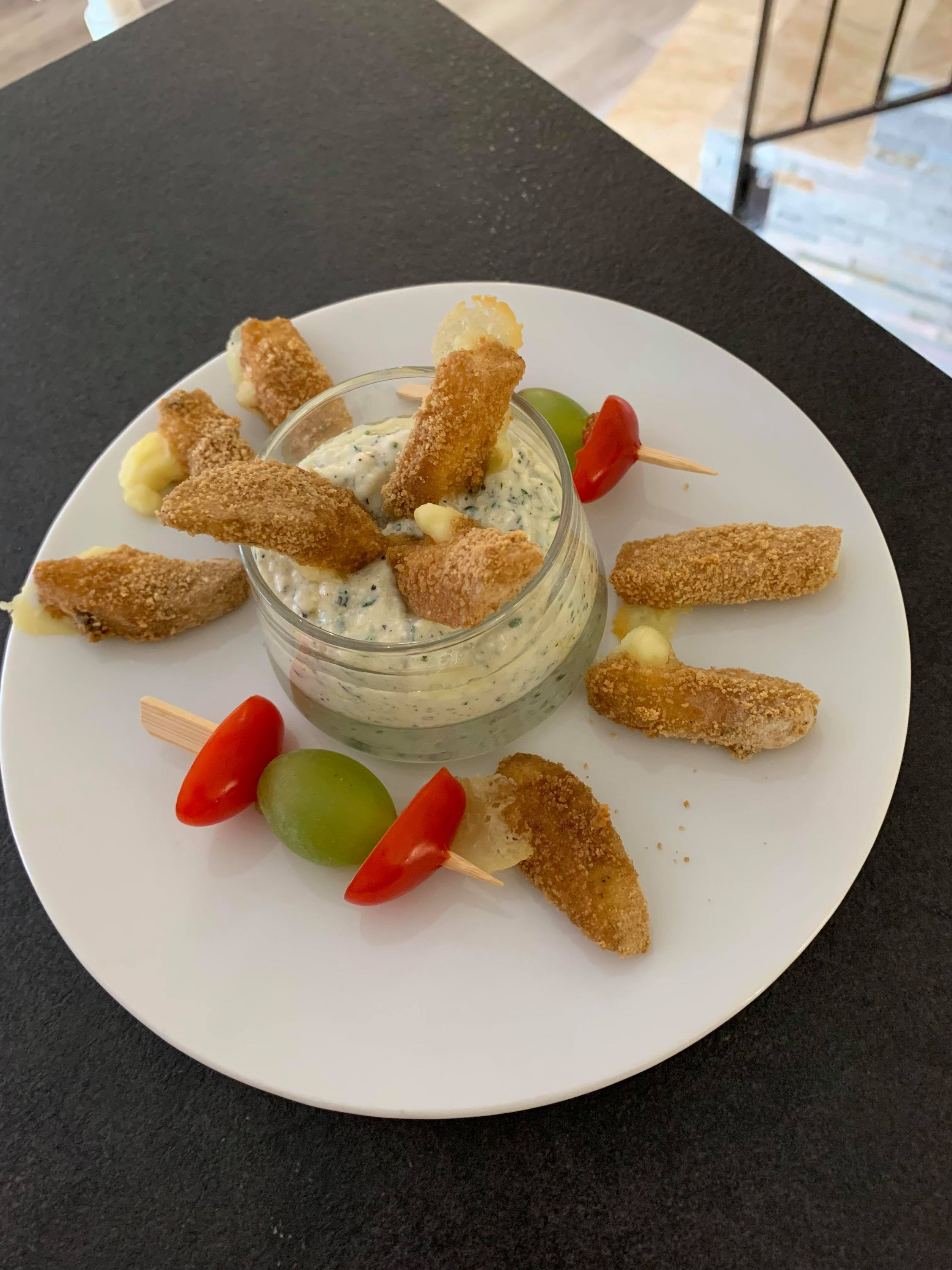 En hvid tallerken med en glas skål med dip, omgivet af panerede snacks og kirsebærtomater.