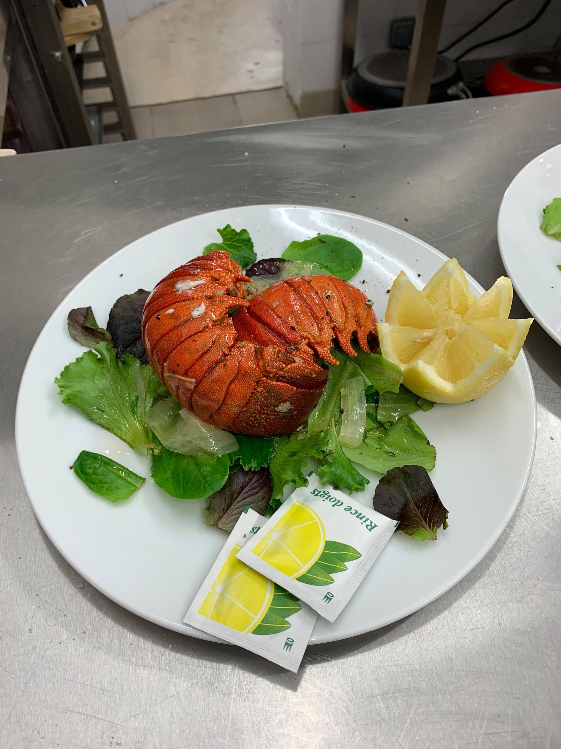 A plate with a cooked lobster, lemon slices, and lettuce on a stainless steel table.