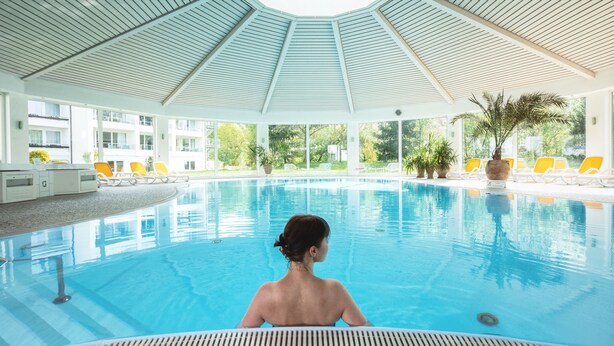 A large indoor swimming pool with clear blue water, surrounded by lounge chairs and potted plants. - Hotel Traube Revital (Wurmlingen)