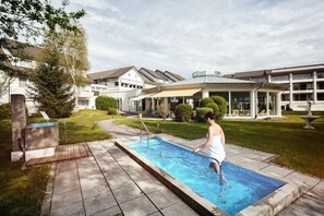 A swimming pool with a wooden deck, surrounded by a well-maintained lawn and a building with a covered patio area. - Hotel Traube Revital (Wurmlingen)
