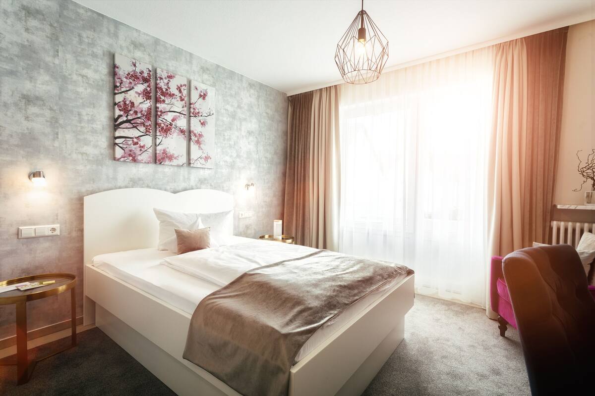 A modern hotel room with a large bed, a bedside table, a lamp, and a window with curtains.