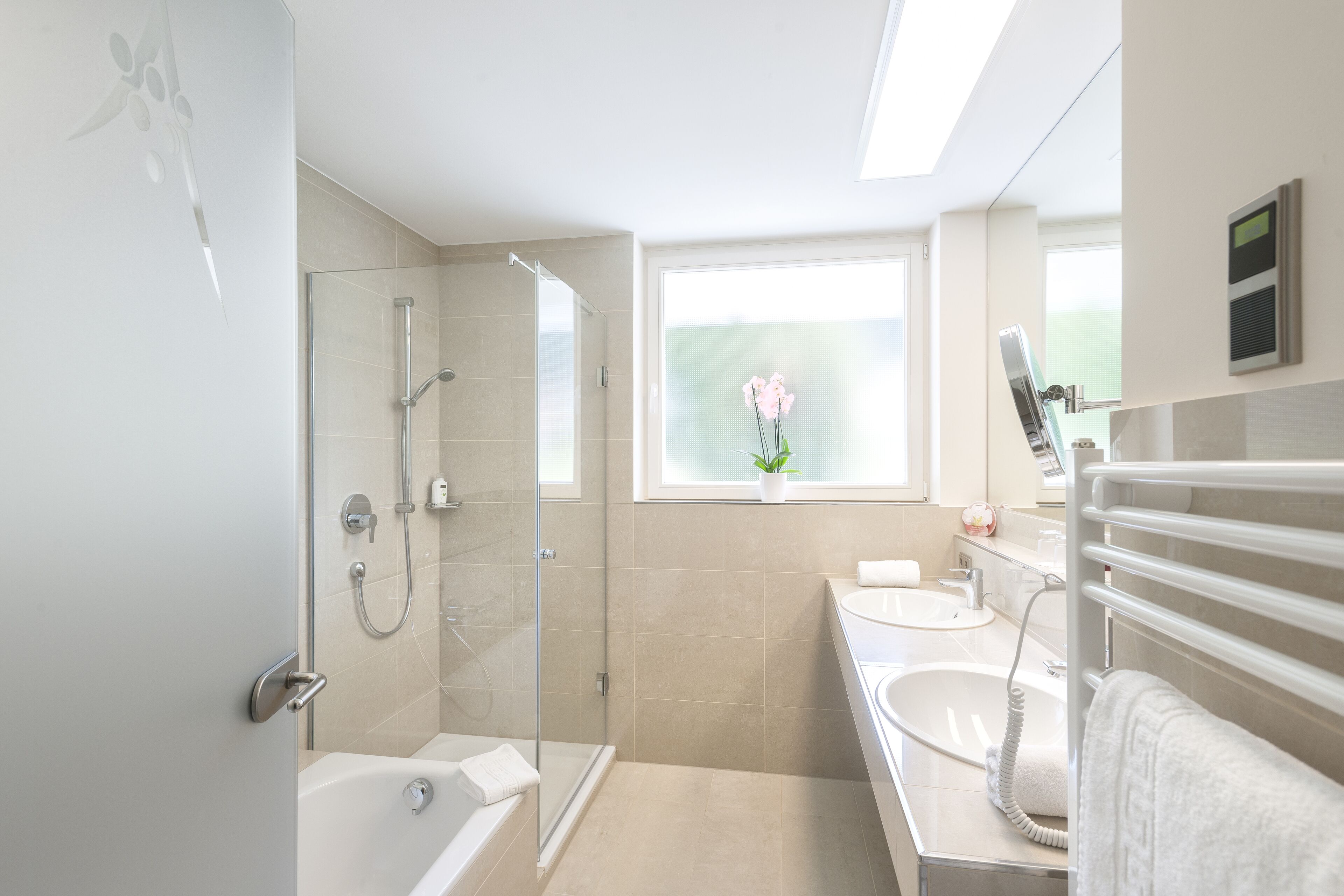 Un baño moderno con ducha de vidrio, bañera, lavamanos y ventana con planta en maceta.