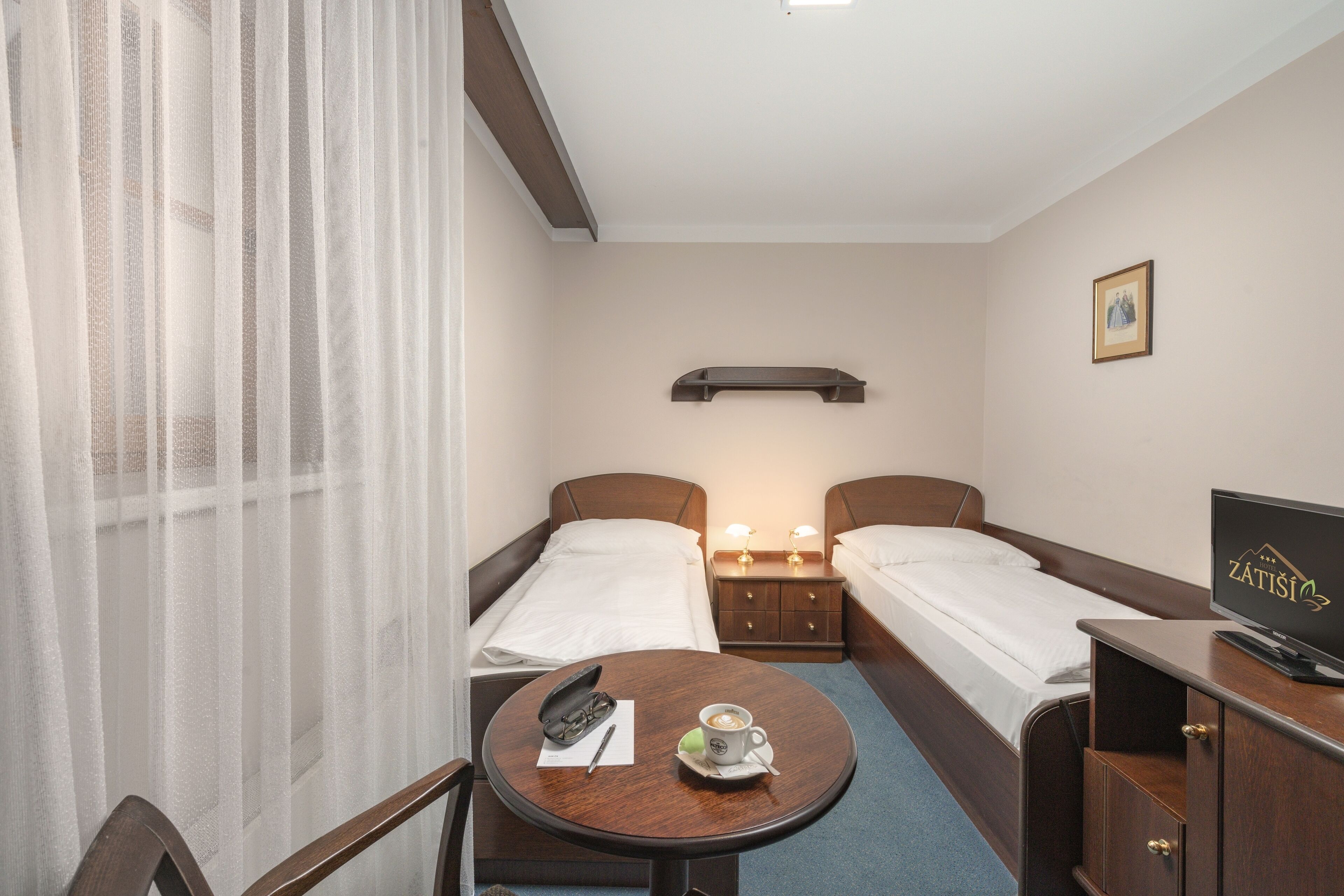 A hotel room with two single beds, a small table with a coffee cup, a TV, and a nightstand with a lamp.
