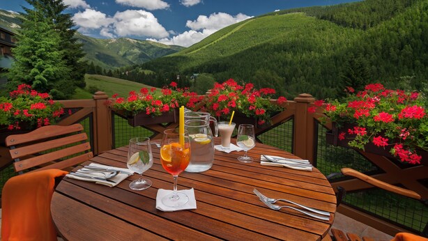 A wooden table set outdoors with a scenic mountain view, a pitcher of iced tea, and red flowers. - Hotel Zátiší (Spindleruv Mlyn)